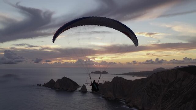 Man paragliding high above coastal cliffs and ocean at sunset, cinematic aerial view of extreme sport flight