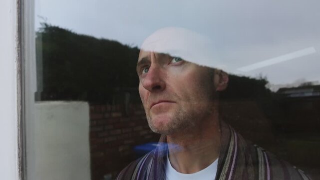 Male midlife crisis. Male actor looks out of house window depressed and bored. Middle aged man wearing a dressing gown. Male mental health crisis, self isolation midlife crisis. 