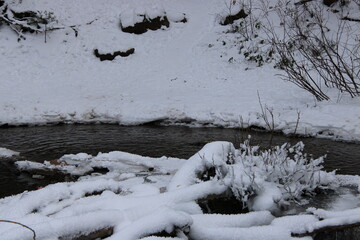 Obraz premium Snow-covered creek bank with flowing dark water, icy edges, and winter vegetation, creating a quiet seasonal landscape with natural contrast and calm atmosphere.