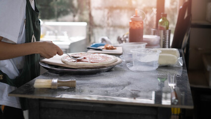 Close-up of chef spreading tomato sauce on raw pizza dough in kitchen, homemade pizza preparation process, artisan cooking concept, fresh ingredients, traditional food making, casual dining lifestyle.