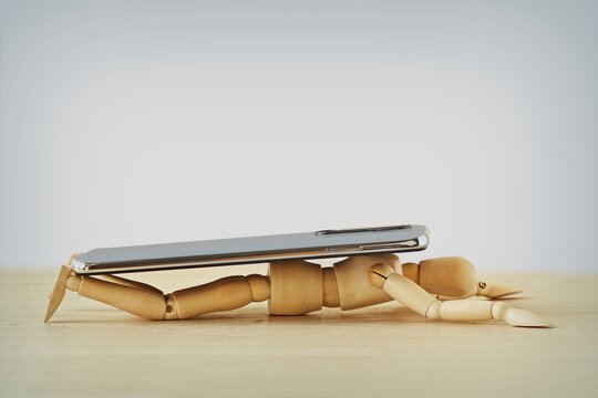 Wooden mannequin crushed under a smartphoe - Technology and mobile devices addiction concept