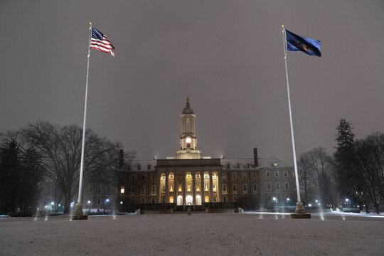 Night view of the historic Old Main building with American and university flags on a snowy campus during the winter season