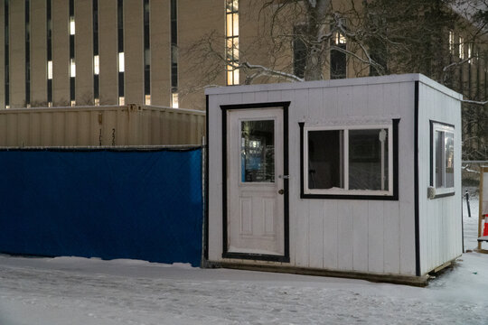 Small White Portable Construction Office Cabin or Security Guard Shack at a Fenced Building Site Illuminated by Night Lights