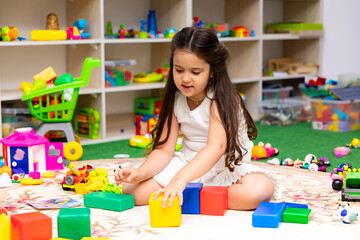 Fototapeta premium Little girl with long dark hair stacking colorful building blocks on a soft rug in a bright playroom filled with toys and storage shelves. Childhood and entertainment concept. High quality photo