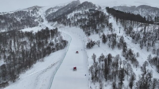 Cinematic drone shot of red snow grooming machines preparing the ski runs for tourists in the Bakuriani mountain resort during the winter season.