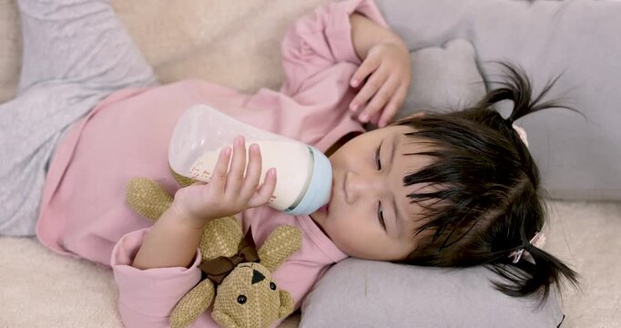 Asian preschool daughter resting on pillow while quietly holding bottle with both hands and hugging plush toy in calm early moment at home representing security, relaxation and self-soothing behavior