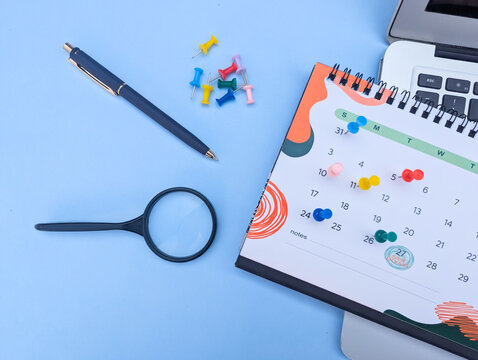 top view of desk calendar with colorful push pins, magnifying glass, and pen on blue background, business planning events management time table concept