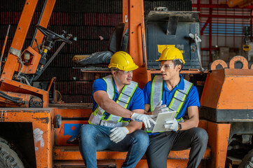 Two warehouse workers wearing hard hats and safety vests sit on a forklift and discuss something while looking at a digital tablet. © BESTIMAGE