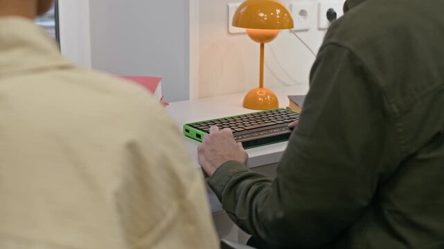 Tilt down shot of mid-aged Caucasian blind man in dark glasses sitting at desk near female assistant and learning how to use computer keyboard with braille display