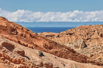 Beautiful landscape of red mountain rocks leading to a blue lake, with clear sky and white clouds. Travel destination in the Tien Shan mountains.