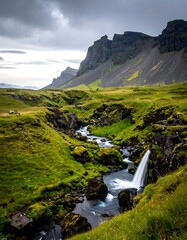 A stream flowing through verdant hills, leading toward imposing, rugged mountain peaks under a cloudy sky