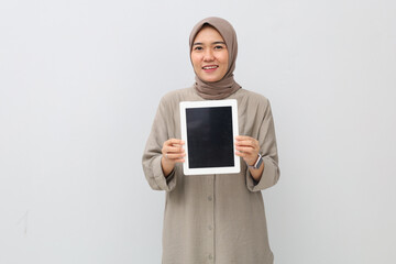 Photo of a stunning young Asian woman in hijab standing on a white background showing a blank digital tablet computer screen to the camera while smiling.