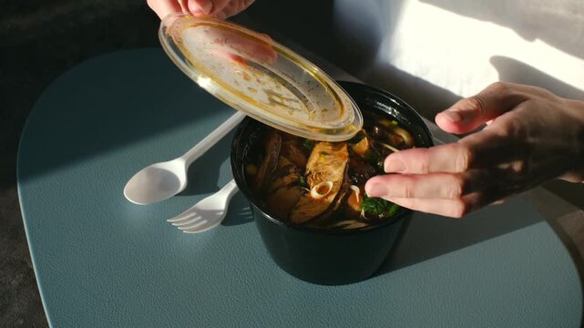 Woman hands opening plastic takeout container of hot asian noodle soup on modern table, with sunlight casting dramatic shadows, revealing ready to eat meal for convenient lunch. Asian noodle soup
