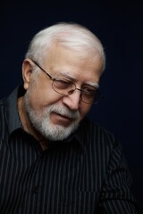 Obraz premium Elderly man with glasses smiles softly while sitting indoors in a dark setting during the evening hours