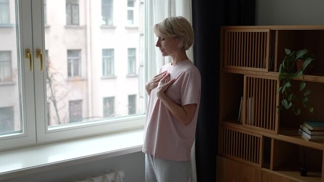 Middle aged woman with closed eyes stands beside window, practicing deep breathing to calm down mind and body, reduce anxiety. Female do relaxation self care rituals for mindfulness and mental health.