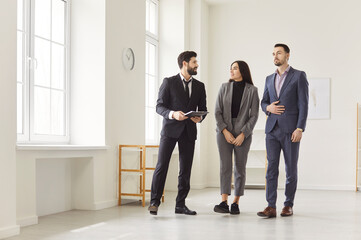 Real estate agent and couple clients in empty room, businesspeople standing, looking around,...