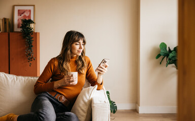 Woman sitting on a sofa, drinking coffee and looking at her smartphone