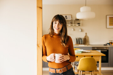 Happy woman holding a hot drink mug, relaxing in her home kitchen interior