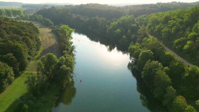 Aerial view of the Kolpa River near Metlika, Slovenia, with a lone canoeist paddling through calm water surrounded by lush green forest, reflected in the river during warm summer light.