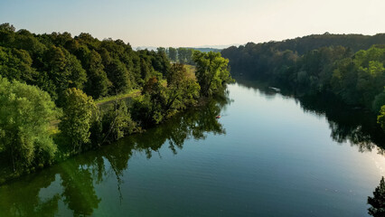 Obraz premium Aerial view of the Kolpa River near Metlika, Slovenia, showing a small river weir, calm green water, surrounding forest, and riverside recreation in a peaceful rural landscape during summer.