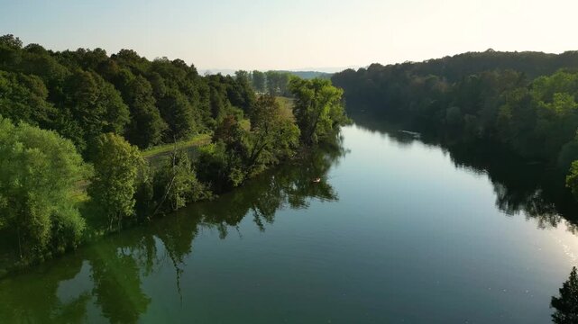 Aerial view of the Kolpa River near Metlika, Slovenia, with a lone canoeist paddling through calm water surrounded by lush green forest, reflected in the river during warm summer light.