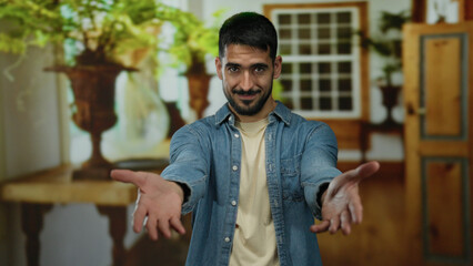 Obraz premium Young man with outstretched arms in a hotel room, wearing a denim shirt, conveying friendliness and warmth in an inviting indoor setting with plants.