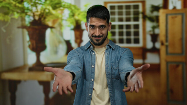 Young man with outstretched arms in a hotel room, wearing a denim shirt, conveying friendliness and warmth in an inviting indoor setting with plants.