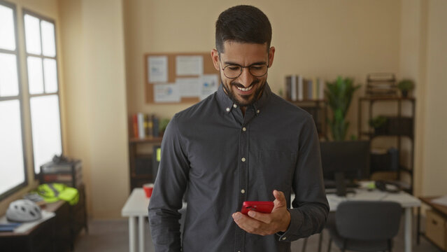 Young man with glasses smiles while using smartphone in modern office featuring bright decor and shelves, creating a professional and relaxed work atmosphere.