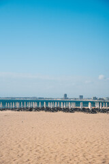 Fototapeta premium Visitors relax on sunbeds with umbrellas at a beach in Bulgaria. The coastline of sunny beach stretches along the water, while buildings rise in the distance. It is daytime and the sky is clear
