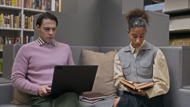 Medium shot of young multiethnic students sitting on couch in comfortable modern library and studying together, working on project using laptop and books