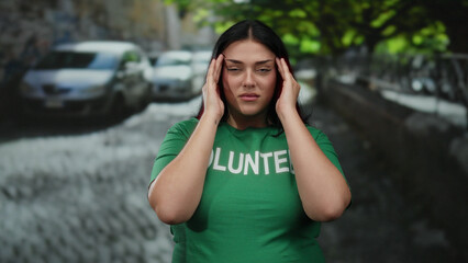 Young woman wearing green volunteer t-shirt feeling headache while standing outdoors on a city...