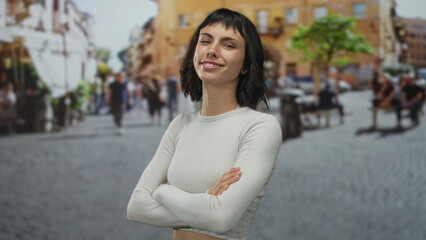 Fototapeta premium Young hispanic woman smiling with crossed arms in busy cobblestone street under bright afternoon sunlight; confidence.