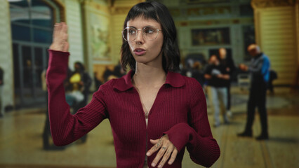 Fototapeta premium Young woman with glasses in maroon sweater extends hands among travellers at airport; confidence and clarity.