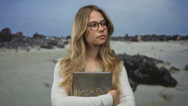 Woman with blonde hair wearing round glasses holds ornate book to chest on rocky shoreline; reflection calm.
