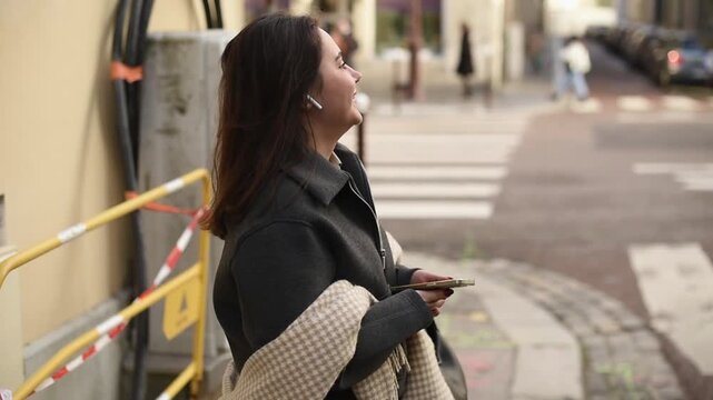 Side view of a woman standing on an urban street wearing wireless earbuds and talking. She is wearing a grey jacket and a patterned scarf draped over her arm.