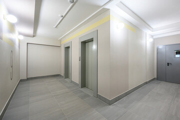 Obraz premium modern hallway with light beige walls, grey tiled floor, recessed ceiling lights, two elevator doors, a metal door on the right, yellow accent stripes, clean and well-lit.