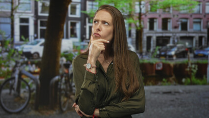 Fototapeta premium Woman hand on chin pensive pose on city street by canal with bicycles and buildings in daylight; curiosity pondering.