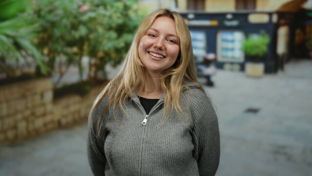 Young blonde woman smiling in city street, wearing grey sweater with outdoor urban background showcasing vibrant greenery and building.