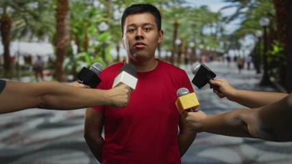Fototapeta premium Man in red shirt with multiple microphones pointed at mouth on street, standing with hands behind back; media anxiety.