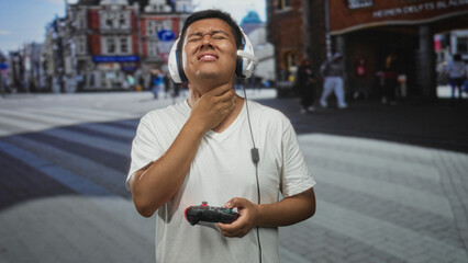 Hispanic young man wearing headset holding game controller with hand on throat on street  discomfort frustration gaming. © Krakenimages.com