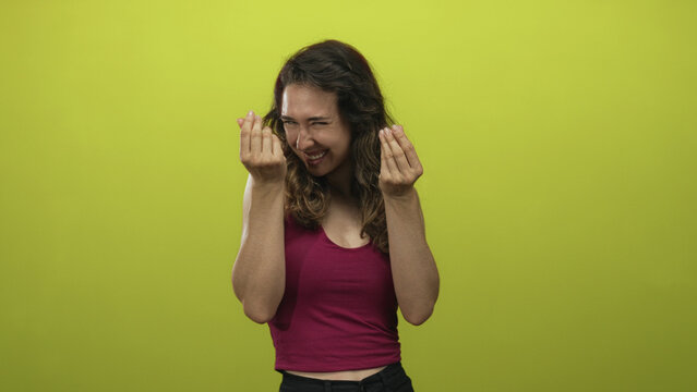 Hispanic woman makes money gesture with fingers against green studio wall; playful wealth desire exuberance.