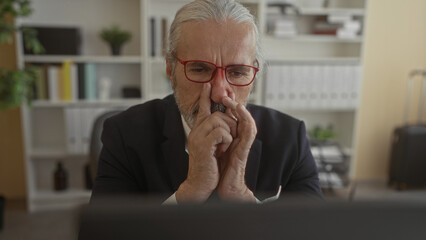 Fototapeta premium Man in suit with hand on chin and fingers touching nose, red glasses, seated at desk in building; thoughtful reflection.