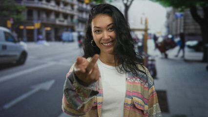 Fototapeta premium Young hispanic woman smiling with hand extended palm up beckoning on a busy city street in daylight; friendly invitation.