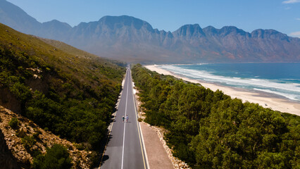 Obraz premium Explore the breathtaking vista along the Kogelbay Beach road in Western Cape, South Africa. Lush greenery meets golden sands under a clear blue sky, inviting travelers and adventure seekers.