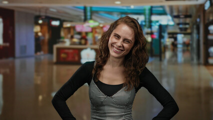 Smiling young woman standing in a mall center with shops in the background, expressing joy in an indoor shopping environment.