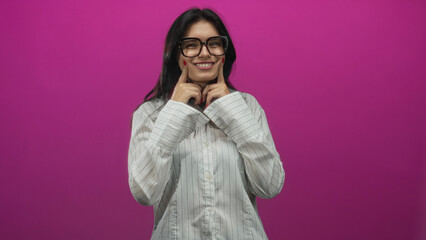 Naklejka premium Young hispanic woman pushes cheeks outward with both index fingers in bright pink studio setting; toothache discomfort.