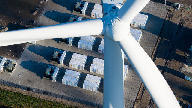 Dronter, Netherlands - 14 January 2026: Aerial view of a colossal white wind turbine overshadowing rows of compact, rectangular energy storage units, casting sharp shadows on the flat landscape.