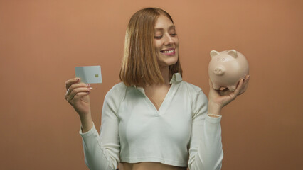 Young blonde woman smiling while holding a credit card and a piggy bank against an isolated brown...