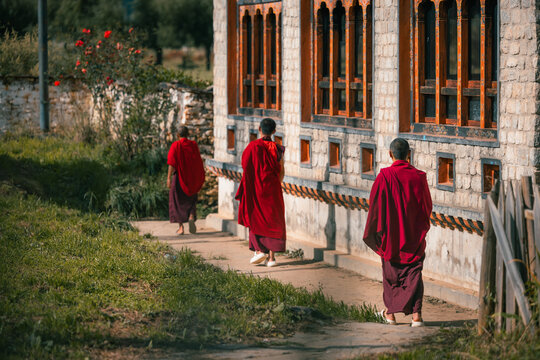 View of monks in vibrant red robes walking along a stone path beside a textured building with carved windows, Jambay Lhakhang, Bumthang, Bhutan.