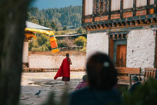 View of a monk in vibrant red robes walks across the stone courtyard of Jambay Lhakhang, framed by traditional architecture and prayer flags, Jakar, Bumthang, Bhutan.